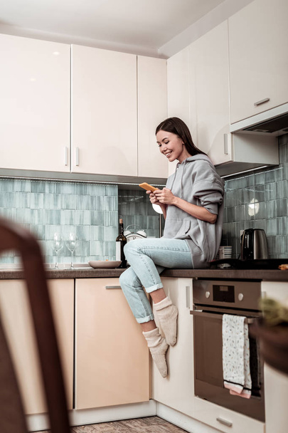 Dark-haired woman wearing jeans and oversize hoodie sitting in kitchen - Foto, immagini