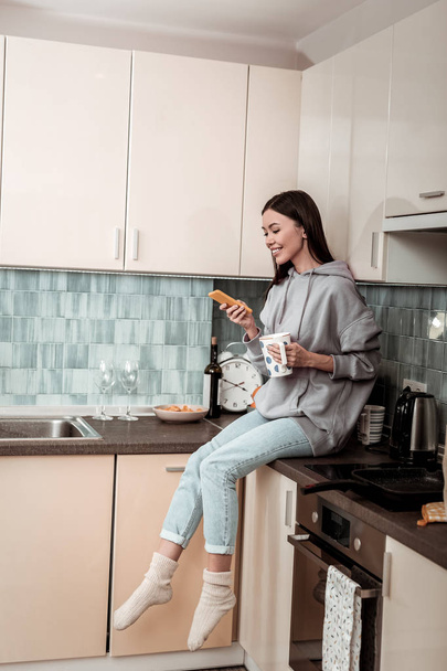 Cheerful woman sitting between cooker and sink reading news - Fotoğraf, Görsel