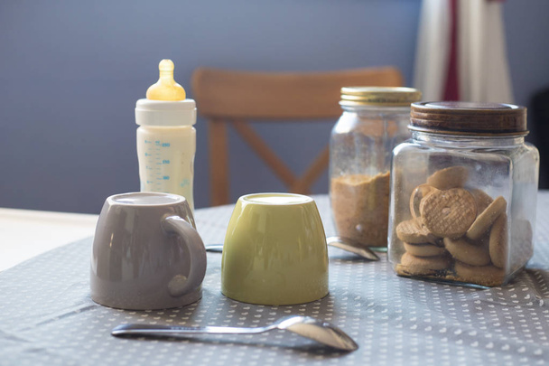 Стол для завтрака в естественном утреннем свете с двумя чашками, печеньем и бутылочкой для кормления ребенка с молоком
 - Фото, изображение