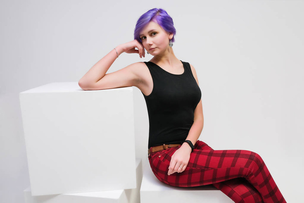 concept beautiful girl with lilac hair on a white background advertises product. She sits in the studio on a white cube right in front of the camera in various poses and shows with her hands. - Fotoğraf, Görsel