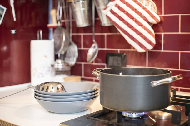 grey cooking pot on gas stove with steam releasing and blue dishes on the side in red burgundy classic kitchen with tools in the background. - Fotó, kép