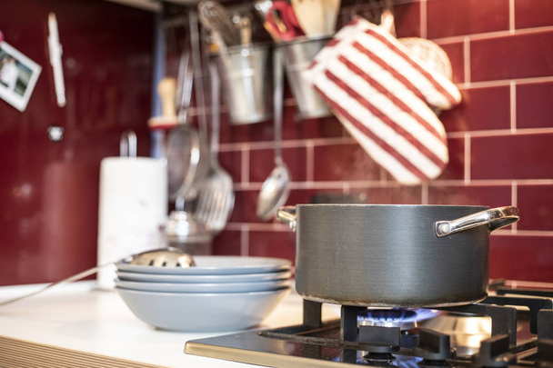 olla de cocción gris en estufa de gas con liberación de vapor y platos azules en el lateral en la cocina clásica roja borgoña con herramientas en el fondo
. - Foto, Imagen