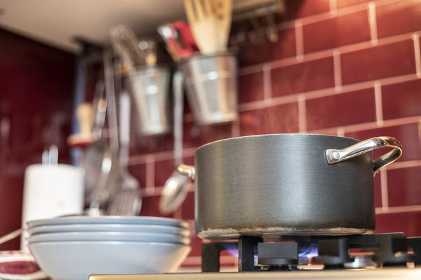 grauer Kochtopf auf Gasherd mit Dampfauslass und blaues Geschirr an der Seite in roter weinroter klassischer Küche mit Werkzeug im Hintergrund. Nahaufnahme. - Foto, Bild