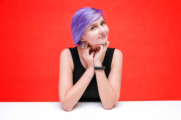 talk to me - Konzeptporträt eines schönen Mädchens mit lila Haaren auf rotem Hintergrund, das am Tisch sitzt und spricht. sie steht direkt vor der Kamera in verschiedenen Posen im Studio. - Foto, Bild