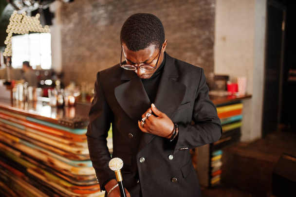 Stylish african american gentleman in elegant black jacket and glasses, holding retro walking stick as cane flask or tippling cane with golden diamond ball handle. Rich fashionable afro man. - Foto, immagini