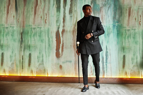 Stylish african american gentleman in elegant black jacket, holding retro walking stick as cane flask or tippling cane with golden diamond ball handle. Rich fashionable afro man against green wall. - Foto, immagini