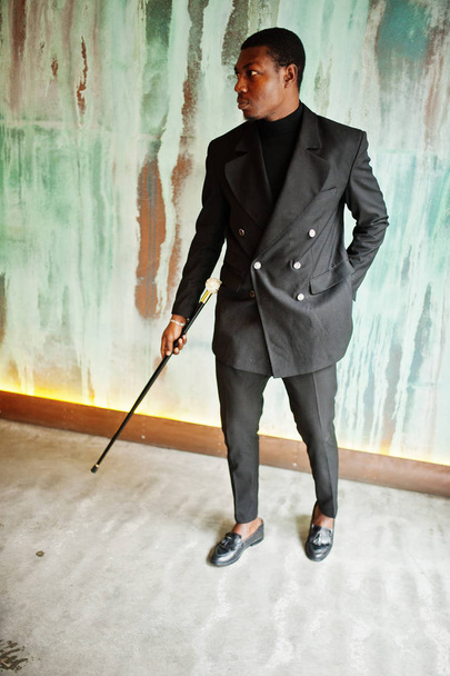 Stylish african american gentleman in elegant black jacket, holding retro walking stick as cane flask or tippling cane with golden diamond ball handle. Rich fashionable afro man against green wall. - Fotoğraf, Görsel
