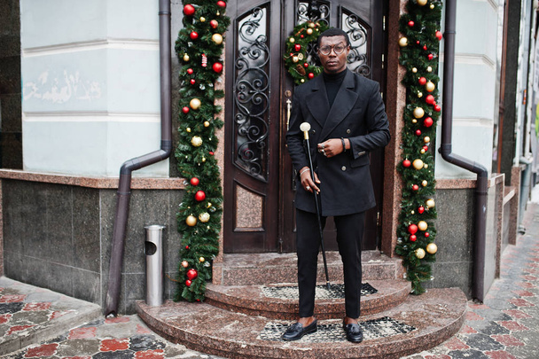 Stylish african american gentleman in elegant black jacket, holding retro walking stick as cane flask or tippling cane with golden diamond ball handle. Rich fashionable afro man against Christmas decorations. - Fotó, kép