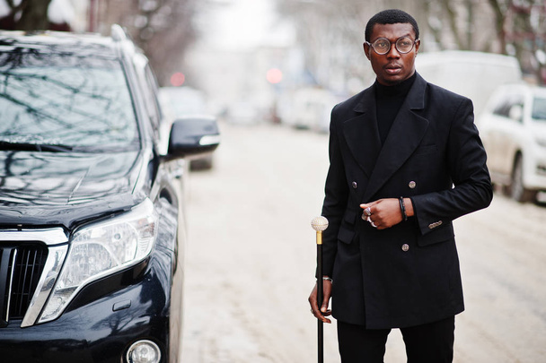 Stylish african american gentleman in elegant black jacket, holding retro walking stick as cane flask or tippling cane with golden diamond ball handle. Rich fashionable afro man against business suv car. - Foto, afbeelding