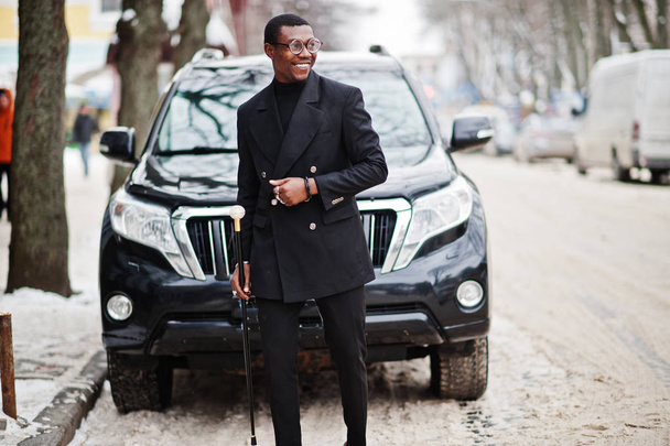 Stylish african american gentleman in elegant black jacket, holding retro walking stick as cane flask or tippling cane with golden diamond ball handle. Rich fashionable afro man against business suv car. - Photo, image