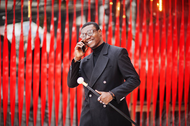 Stylish african american gentleman in elegant black jacket, holding retro walking stick as cane flask or tippling cane. Rich fashionable afro man against red wall speaking on mobile phone. - Fotó, kép