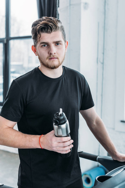 handsome young man holding sports bottle and looking at camera in gym - Фото, зображення