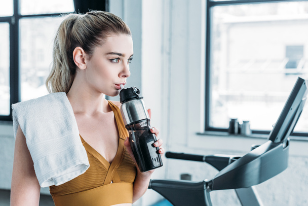 beautiful sportswoman with towel on shoulder drinking water from sports bottle in gym - Photo, Image