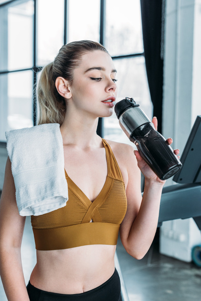 sporty young woman with towel on shoulder drinking water from sports bottle in gym - Photo, Image