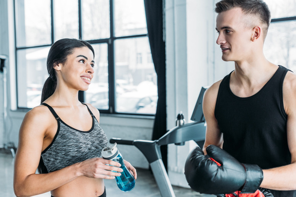 young man with boxing gloves and african american woman with sports bottle smiling each other in gym - Фото, зображення