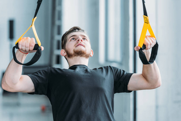 handsome muscular young man exercising with resistance bands and looking up in gym - Valokuva, kuva