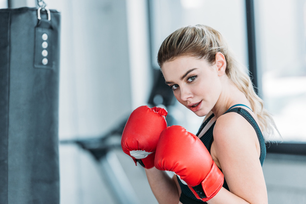 attractive young sportswoman in boxing gloves looking at camera in gym - 写真・画像