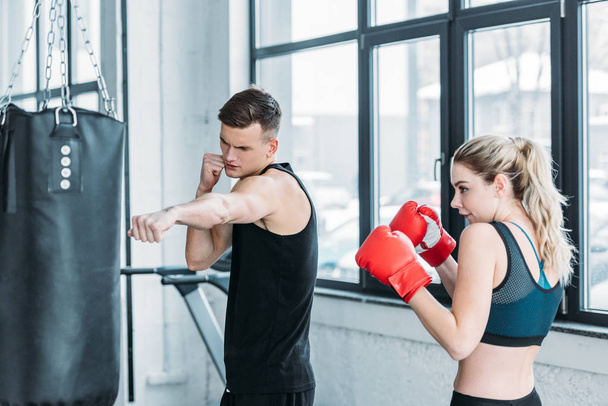 izmos férfi oktató és fiatal nő Boxkesztyű képzés boxzsák a tornateremben - Fotó, kép