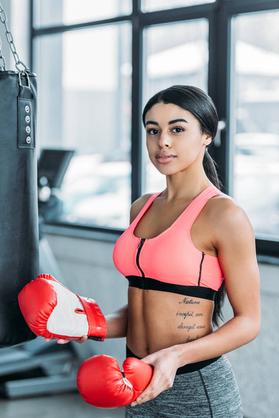 young african american sportswoman holding boxing gloves and looking at camera in gym - Фото, зображення