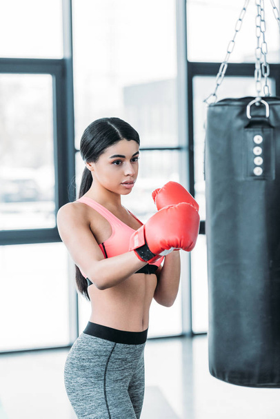 concentrated african american woman in boxing gloves training with punching bag in gym   - Photo, Image