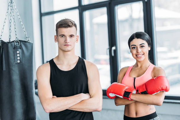 male trainer and young african american woman in boxing gloves standing together and looking at camera in gym - 写真・画像