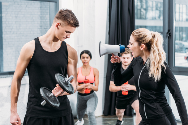 sportswoman with megaphone yelling at young man training with dumbbell in gym - Zdjęcie, obraz