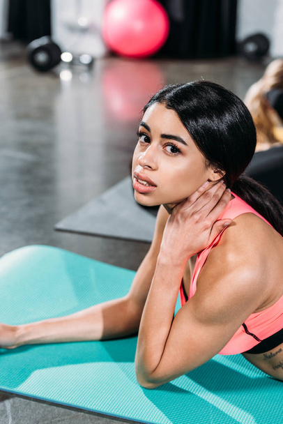 young african american woman touching neck and looking at camera while lying on yoga mat in gym - Fotó, kép