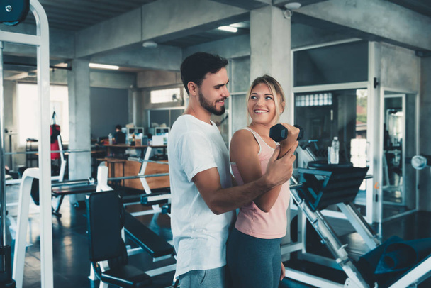 Sporty couple exercising dumbbell weights lifting in fitness gym., Portrait of attractive young couple are practicing workout in training class., Beautiful woman with trainer exercise weights lifting. - Фото, изображение