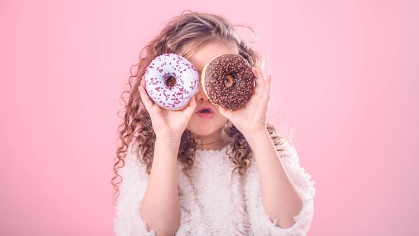 Portrait of a little surprised girl with curly hair, and two mouth-watering donuts in her hands, closes her eyes with donuts, on a pink background - Photo, Image