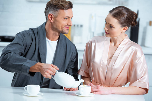 selective focus of beautiful adult couple in robes having tea in kitchen - Foto, Imagem
