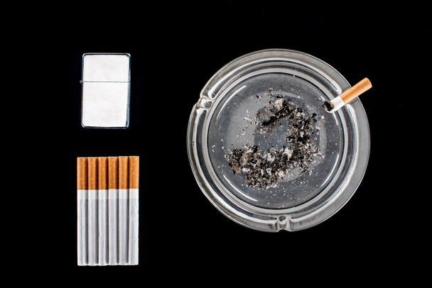 Colocación plana de cenicero de vidrio, encendedor de cigarrillos y cigarrillos aislados en negro
 - Foto, Imagen