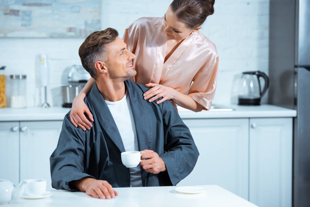 Женщина в халате обнимает красивого мужчину с чашкой чая на кухне
 - Фото, изображение
