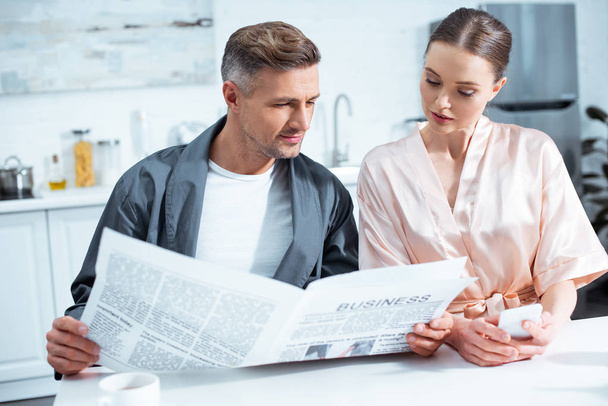 selective focus of couple in robes reading business newspaper in kitchen - Фото, изображение