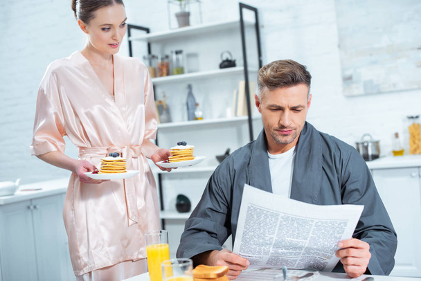 woman in robe holding plates with pancakes while man reading newspaper during breakfast in kitchen - Фото, зображення