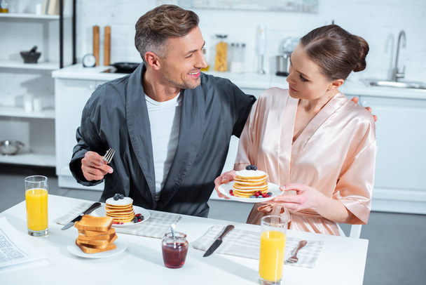 selective focus of adult couple in robes during breakfast with pancakes and orange juice in kitchen - Zdjęcie, obraz