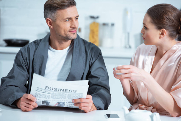 man in robe reading business newspaper while woman drinking tea during breakfast in kitchen - Photo, Image