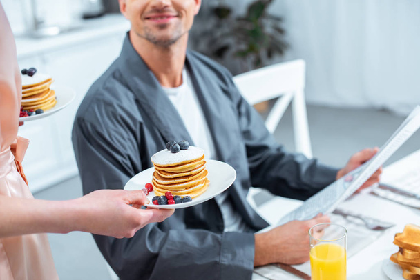 cropped view of woman holding plates with pancakes while man reading newspaper during breakfast in kitchen - Photo, image
