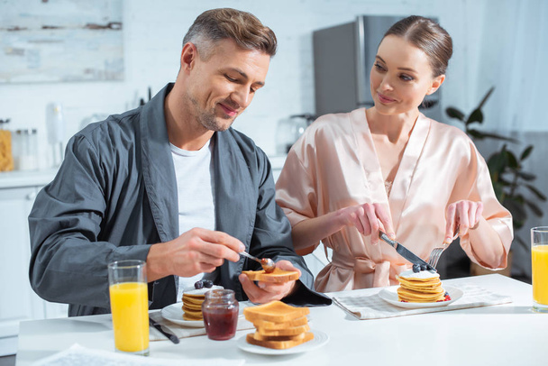 selective focus of husband and wife in robes during breakfast with pancakes and orange juice in kitchen - Φωτογραφία, εικόνα