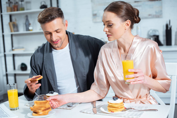 enfoque selectivo del marido y la esposa en túnicas durante el desayuno con panqueques y jugo de naranja en la cocina
 - Foto, Imagen