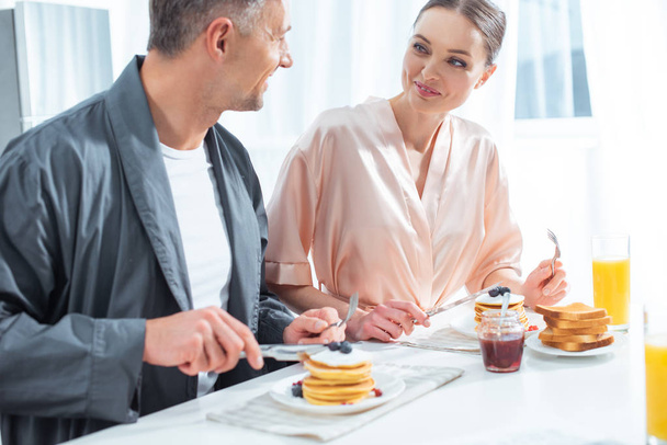 selective focus of beautiful couple in robes during breakfast with pancakes and orange juice in kitchen - Foto, Imagem