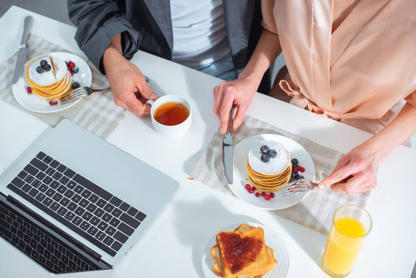 cropped view of adult couple sitting at table with laptop while having breakfast in morning - Foto, Imagen
