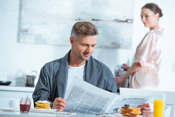 man reading newspaper during breakfast while woman cooking on background  - Fotó, kép