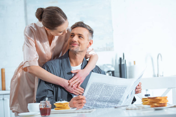 woman in robe embracing handsome man with newspaper during breakfast in kitchen - Фото, изображение