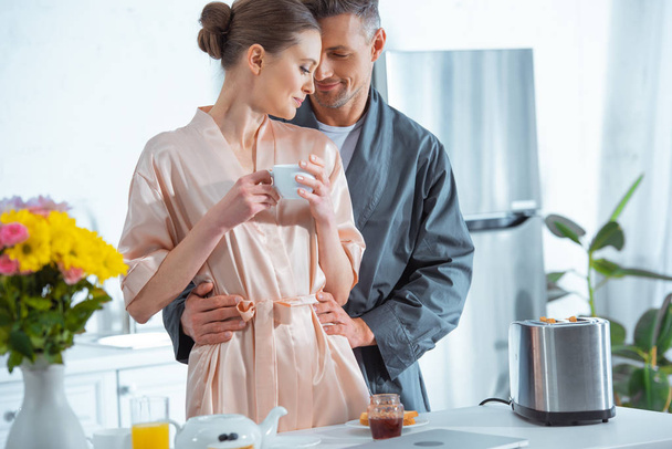 handsome man in robe embracing beautiful woman with cup of tea in kitchen - Фото, изображение