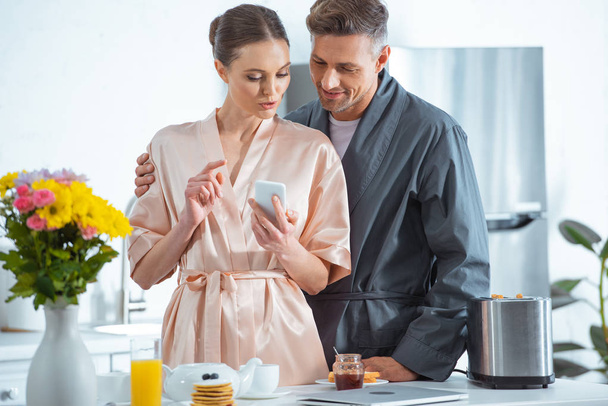 beautiful adult couple in robes using smartphone during breakfast in kitchen - Foto, Imagem