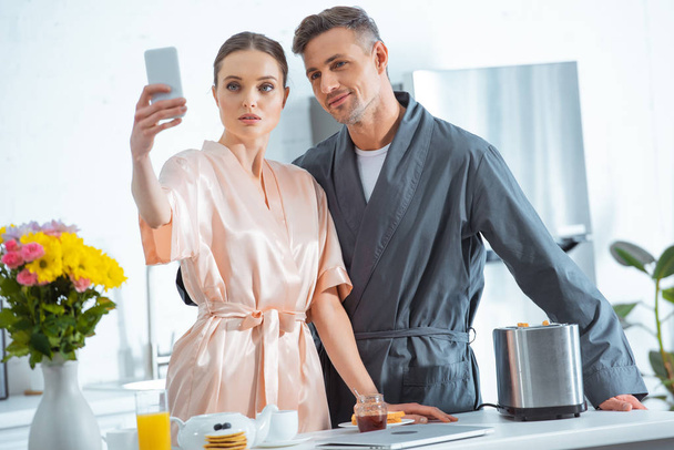 beautiful adult couple in robes taking selfie on smartphone during breakfast in kitchen - 写真・画像