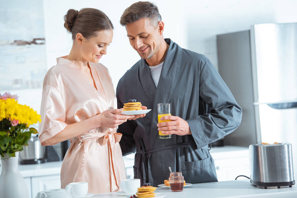 beautiful smiling couple in robes with orange juice and pancakes during breakfast in kitchen - 写真・画像