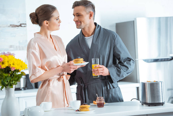 beautiful couple in robes with orange juice and pancakes during breakfast in kitchen - 写真・画像