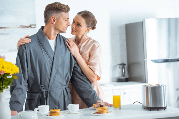 beautiful woman in robe embracing handsome man during breakfast in kitchen - Photo, Image