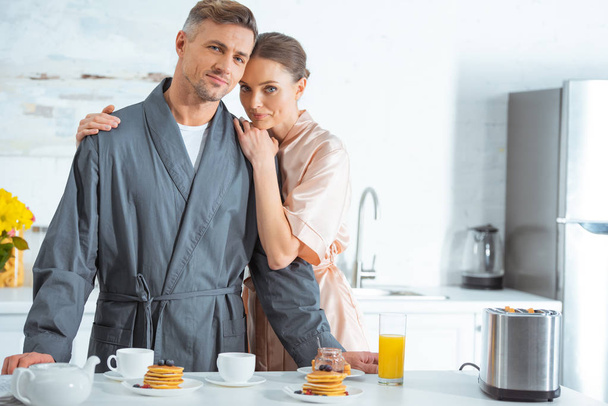 selective focus of beautiful woman in robe embracing man during breakfast in kitchen - Foto, afbeelding
